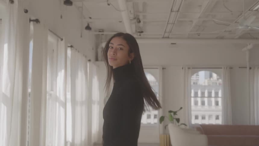 A woman in a black turtleneck walks confidently across a bright elegant room with large windows looking back at the camera with a smile