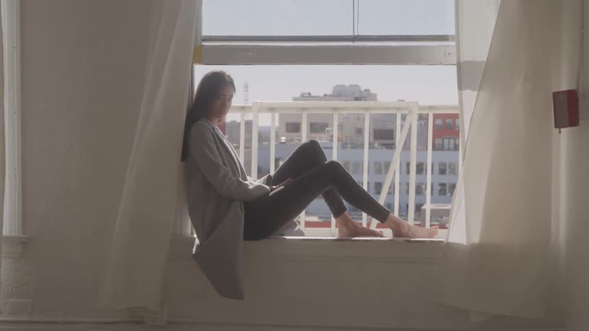 A woman with long dark hair wearing a grey cardigan and dark leggings sits barefoot and quietly on a wide window ledge