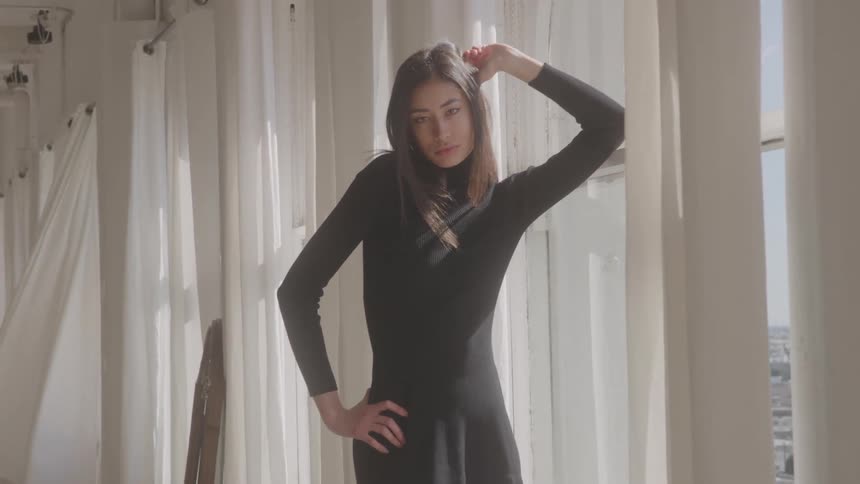 Young female model wearing a black dress posing next to a window indoors during day time