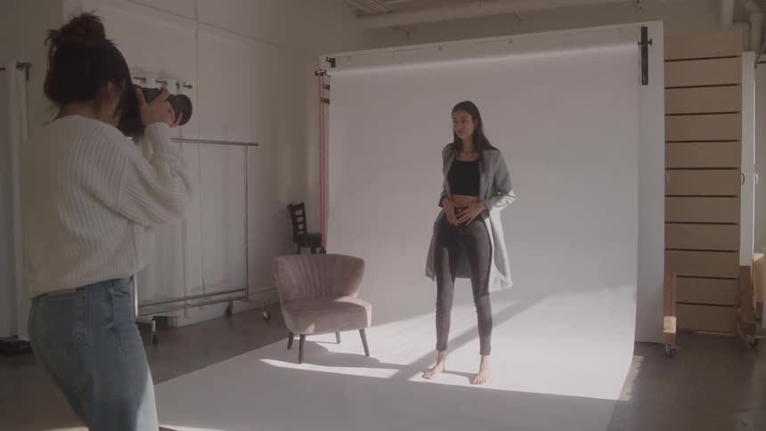 A female photographer captures a beautiful woman posing in a grey blazer and black jeans against a white backdrop in a sunlit photo studio