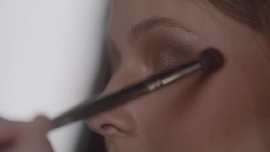 A person calmly closes their eyes while a makeup brush blends warm shadow across her eyelid