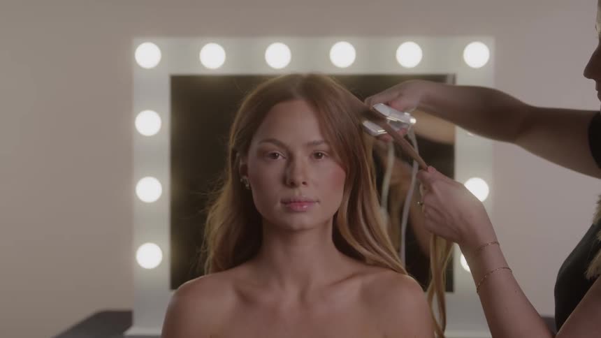 A young woman model with long wavy light brown hair sits facing the camera with bare shoulders as a stylist woman uses a hair straightener on a section of her hair in front of a makeup mirror with globe lights in a studio