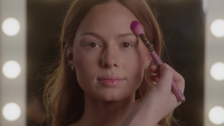 A closeup view shows a makeup artists hand applying pink blush with a brush to the forehead of a young woman with long light brown hair who looks calmly at the camera in a bright vanity mirror setting