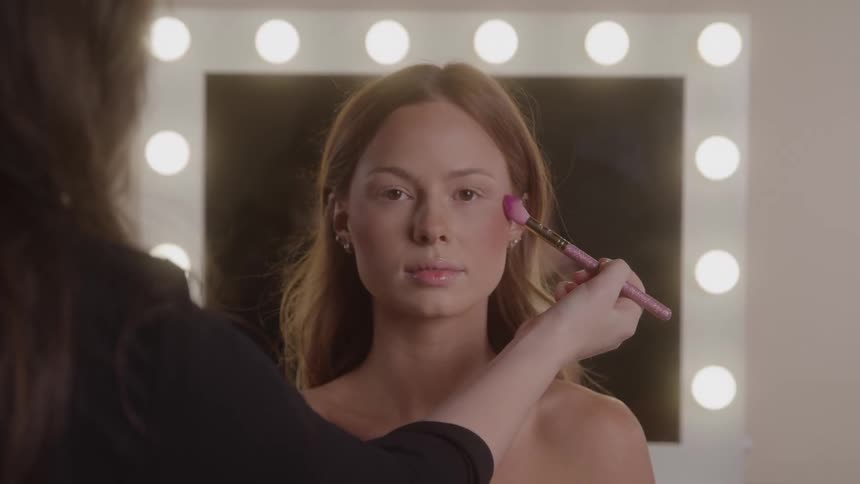 Young female model sitting down while a female makeup artist applies makeup on her face with a brush indoors