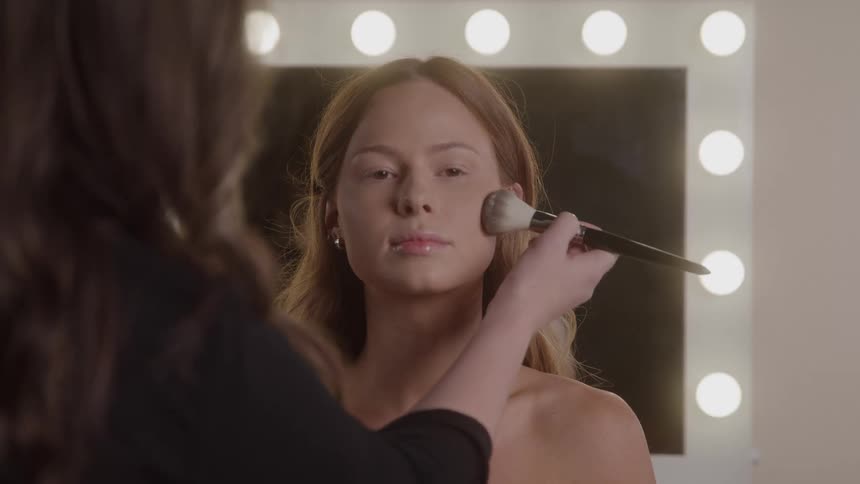A young blondehaired woman sits inside a salon before a large mirror with bright lights at the sides remaining still as another woman applies makeup to her face with a brush