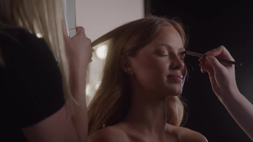 A woman with closed eyes and long hair is simultaneously attended to by two people one applying makeup near her right eye with a brush and the other adjusting her hair on the left side