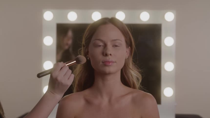 Woman has makeup applied with a brush in front of an illuminated mirror