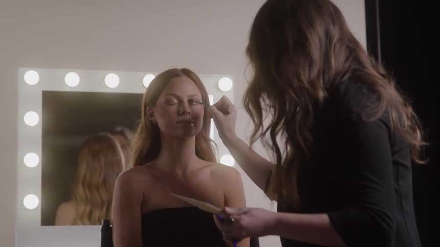 Woman applies makeup to another woman in front of a lit mirror