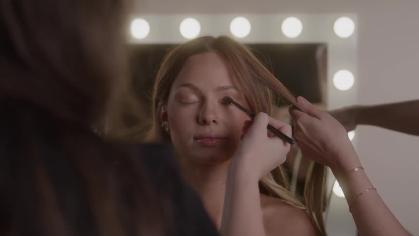 A woman with closed eyes is attended to by two people one applying makeup to her eye with a small brush and the other styling her hair with a flat iron