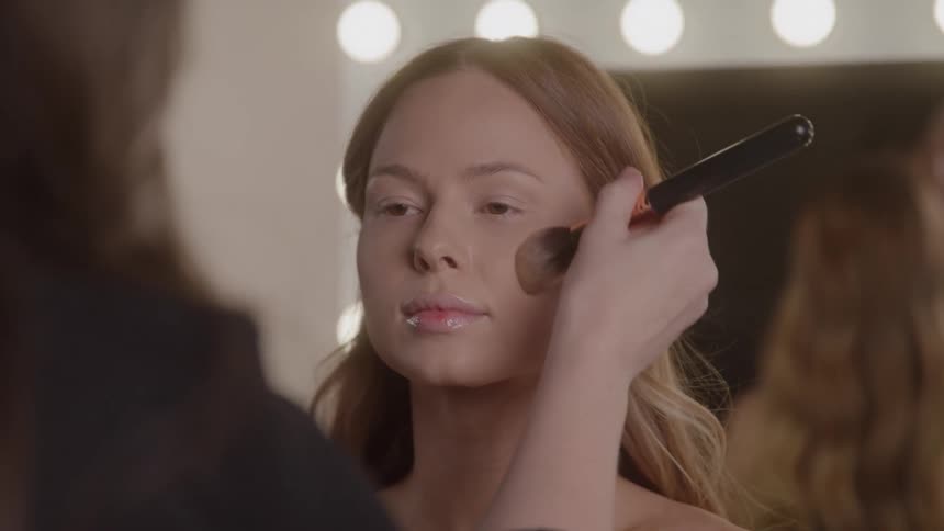 A closeup shot shows a person applying makeup to the cheek of a woman with a large brush while the woman looks straight ahead with her eyes open