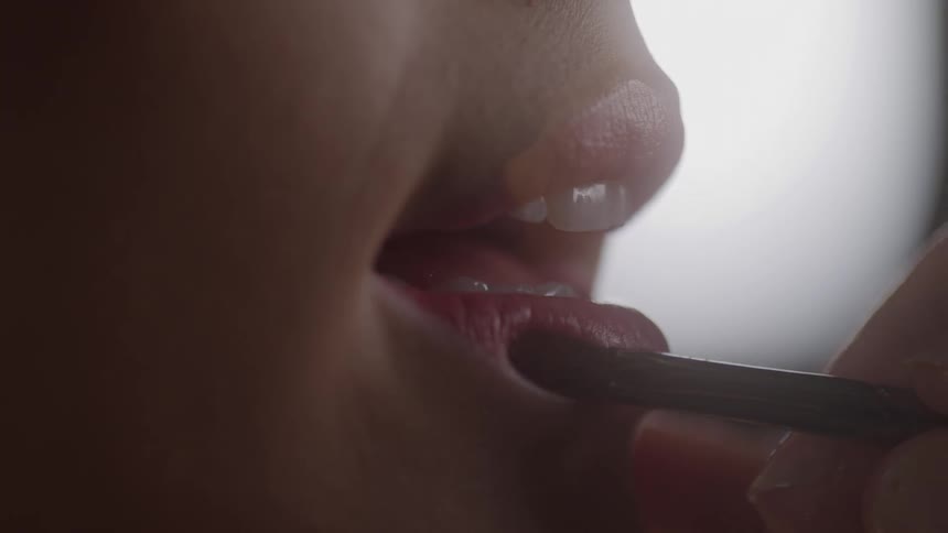 A close view shows a womans lips being delicately touched with a lip brush as she applies dark red lipstick in a makeup application scenario