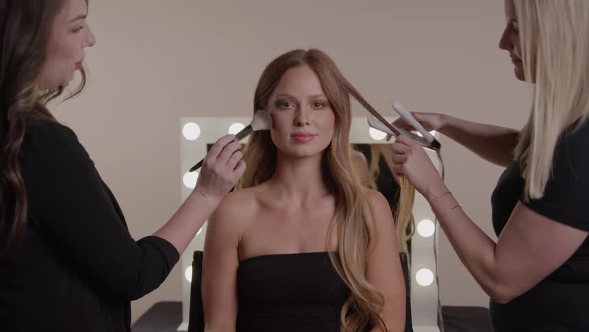 A young woman in a black dress sits at midday in a salon as two other women style her hair