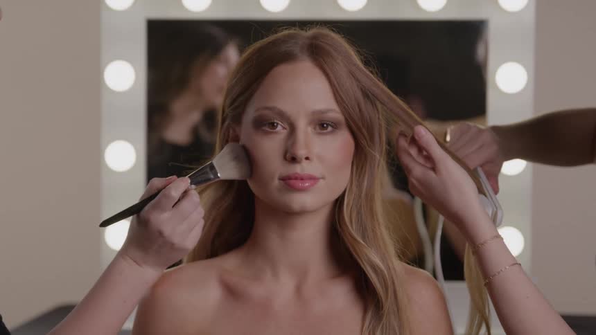 A woman with long hair sits still and looks directly at the camera while two different people simultaneously work on her one applying face powder with a large brush to her left cheek and the other styling her hair with a flat iron on the right side