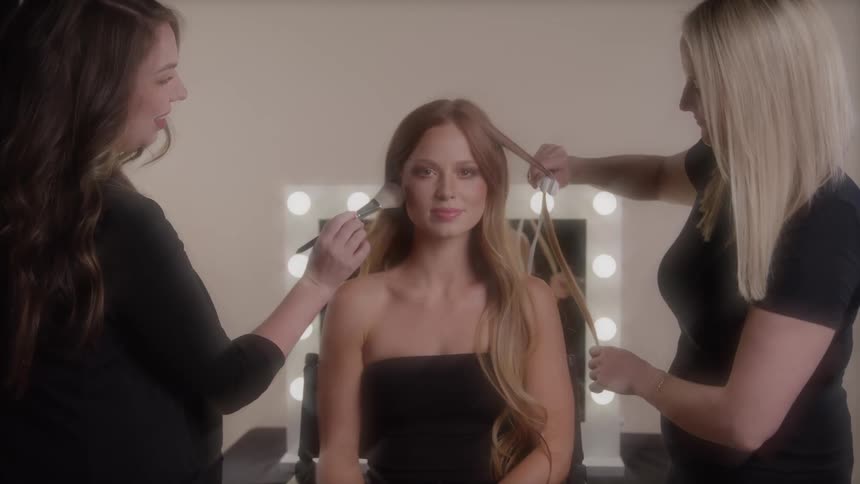 Woman being styled and made up by two professionals in front of a lighted mirror