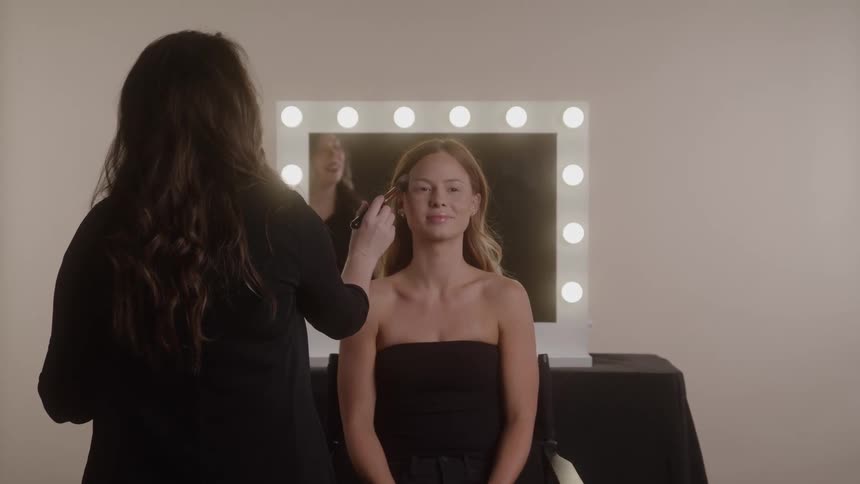 Woman being made up by a professional in front of a lighted mirror in a studio setting