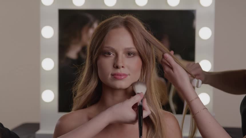 Woman being styled and made up by a team in front of a lighted mirror in a dressing room