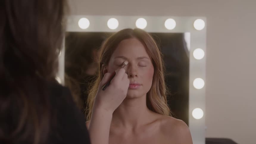 Makeup artist applying eyeshadow to a woman seated in front of a lighted mirror
