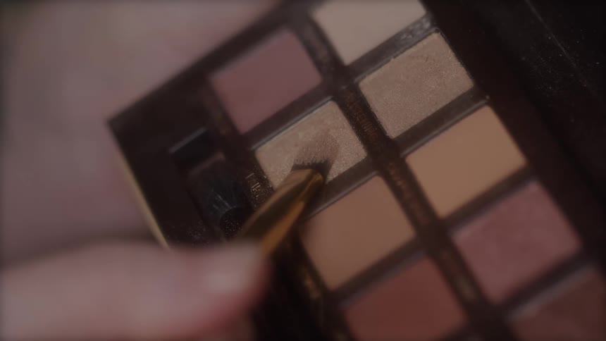 A very closeup shot shows a small angled brush being dipped into a square pan of shimmer eyeshadow within a makeup palette that contains multiple squares of earthtoned powders