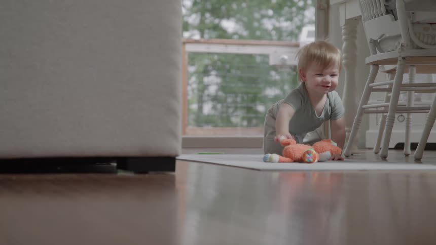 A young child with a bright open smile sits on a mat on a shiny wooden floor next to an orange stuffed animal