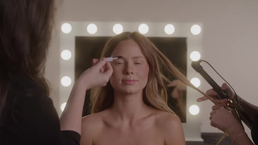 A woman gets her makeup and hair styled at a vanity mirror