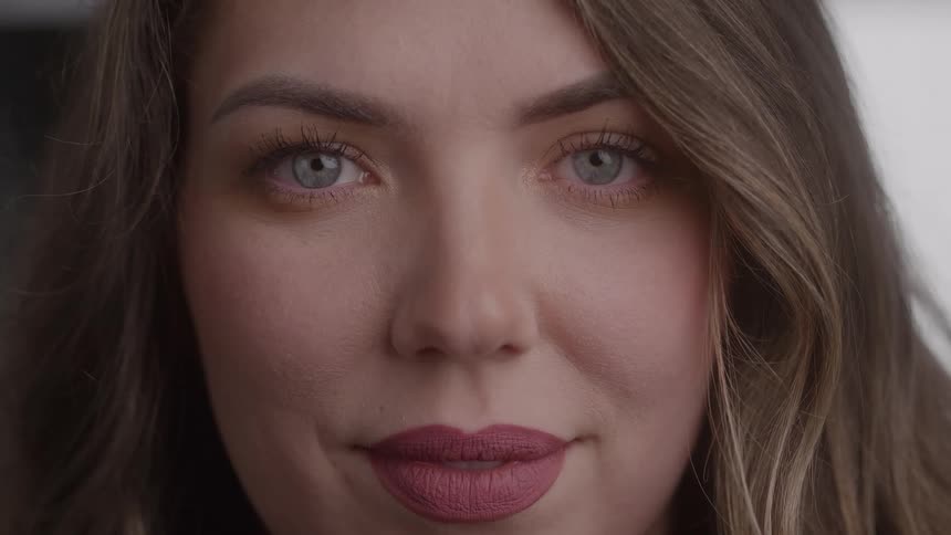 A closeup view of a womans face shows her opening her blue eyes and looking directly at the camera while wearing mauve lipstick and having long brown hair