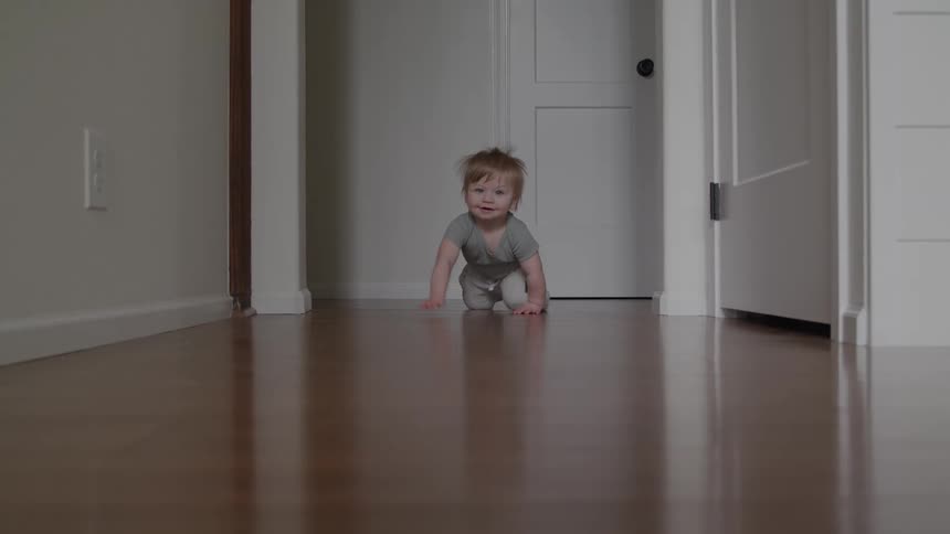 A happy fairhaired baby wearing a grey sleeper is seen crawling directly toward the camera down a shiny wood hallway floor in a modern bright home interior