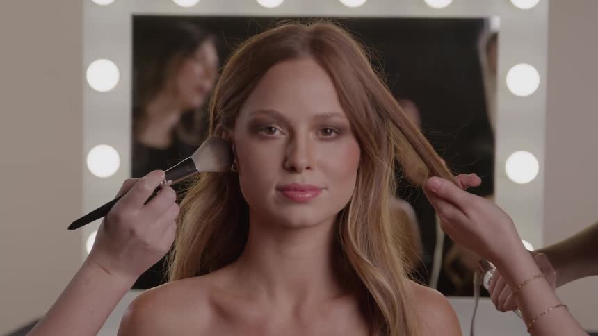 Woman getting makeup and hair done simultaneously in front of an illuminated mirror