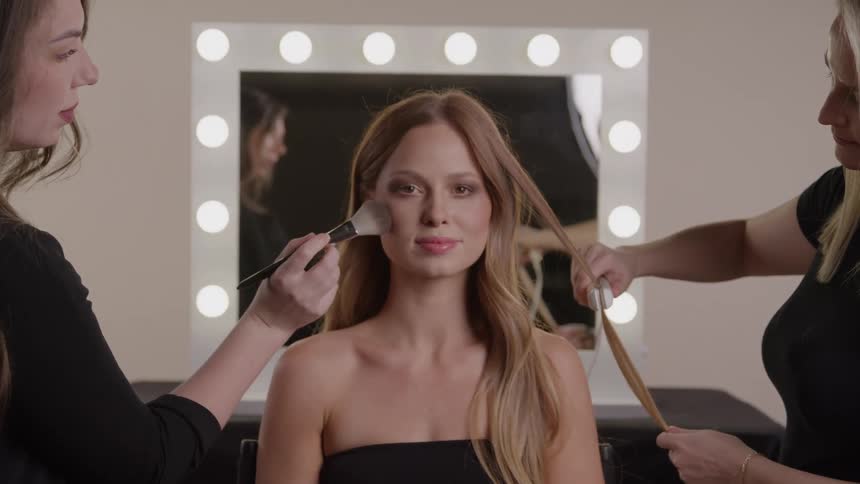 Woman being styled and having makeup applied by two professionals in front of an illuminated mirror