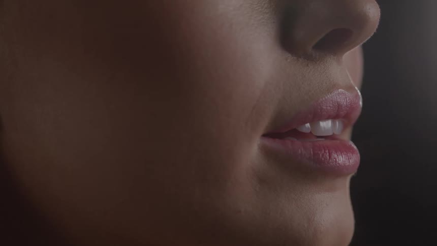 An extreme closeup side profile shot shows the lower half of a persons face as a brush finishes applying a shiny pink lip product to the lips