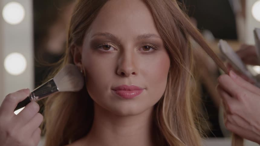 A woman with long hair and a finished makeup look sits centrally and looks directly at the camera while a person on the left applies face makeup with a large brush and another person on the right styles her hair with a flat iron
