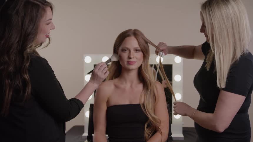 Woman having makeup applied and hair styled by two professionals in a lit dressing room