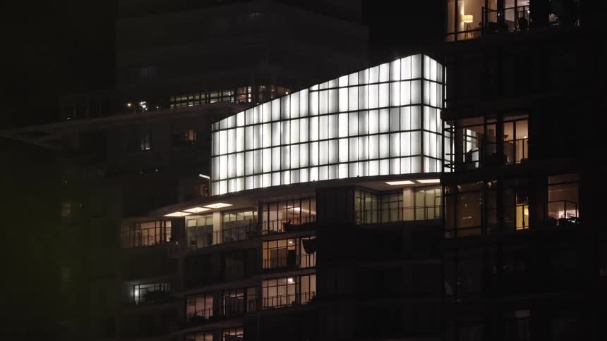 A modern apartment building is shown at night with a brightly illuminated curved glass facade above several floors of dark apartments with warm lights visible inside