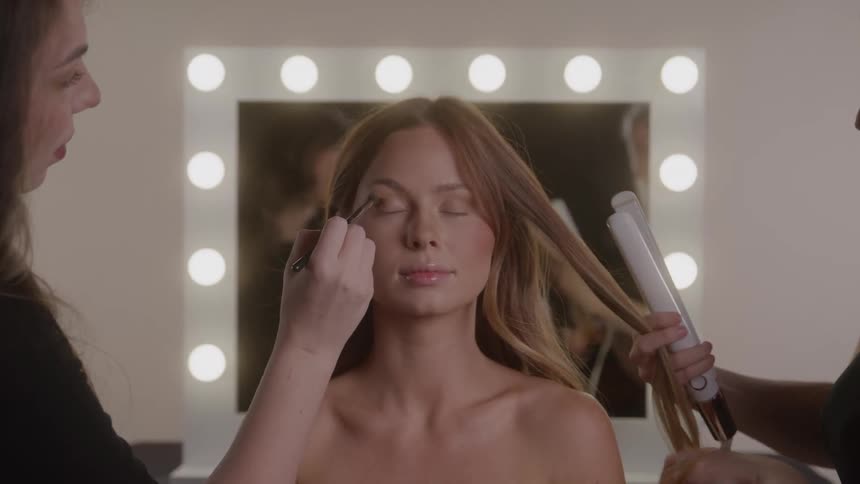 Woman getting her makeup and hair done by two professionals in front of a lighted mirror eyes closed during preparation