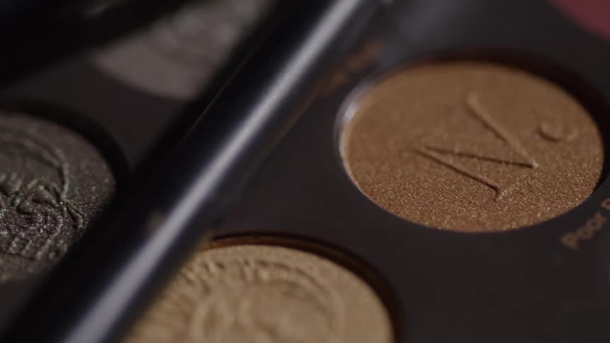 A close view moves across a makeup palette showing various circular eyeshadow colors with embossed designs including shimmering metallic browns grays and a matte reddish shade