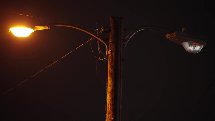 A utility pole holds two contrasting streetlights against a dark night sky with the left light emitting a warm orange glow and the right light appearing unlit or a cooler white suggesting an upgrade or malfunction
