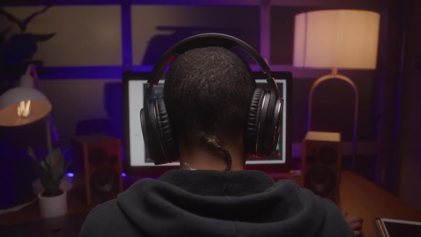 Headphoned person works at a glowing computer in a cozy dim studio with speakers and lamps