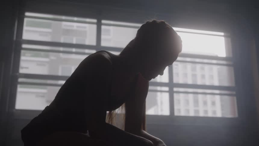 A silhouetted person sits with head bowed in front of a window