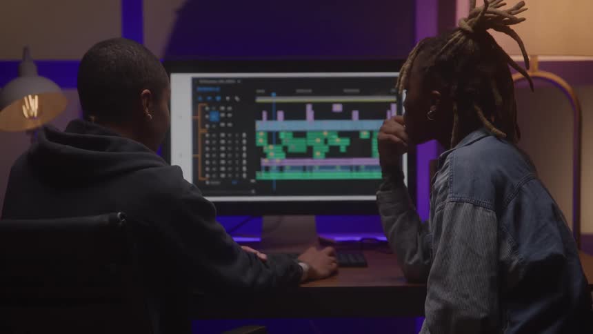 Two people watch and edit a video project on a large monitor in a softly lit room