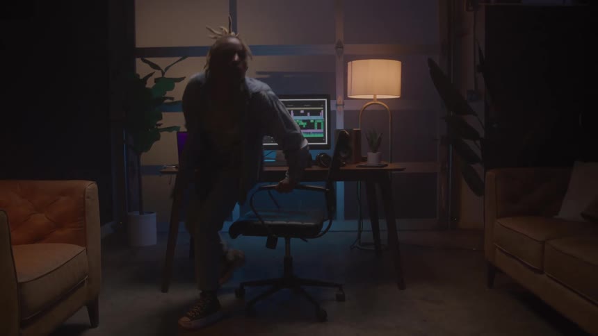 A man with dreadlocks wearing headphones and casual clothes works on a computer with video editing software in a dark stylish home office before suddenly standing and walking away leaving glowing screens and lamps on