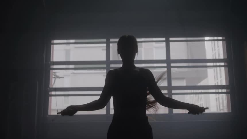 The dark silhouette of a person with long hair is seen jumping rope in a large dimly lit room with a bright gridpatterned window in the background