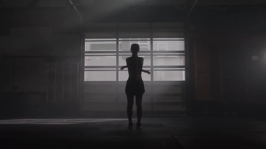 Woman standing in a dim gym stretches in front of a large illuminated window
