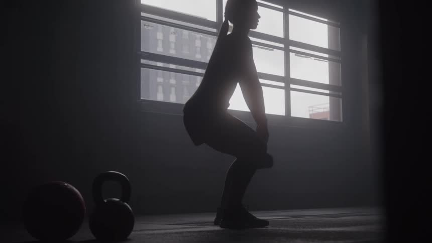 A silhouetted woman wearing shorts and a tank top performs squats while holding a single kettlebell in front of a large brightly lit window in a dark gym
