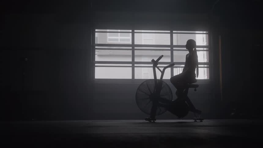 Silhouetted athlete pauses on an air bike in a dim gym head bowed in exhausted recovery