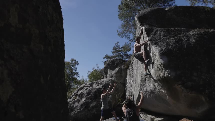 A young man climbing a large rock with the guidance of friends outdoors during daytime