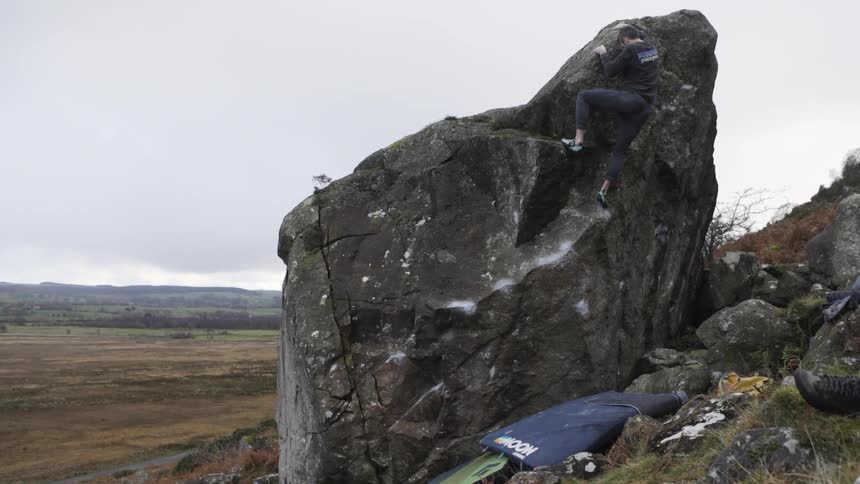 Person climbs a large rock outdoors amid a vast natural landscape