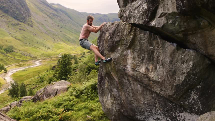 Shirtless man climbs a large rock amid a lush landscape of mountains and valleys