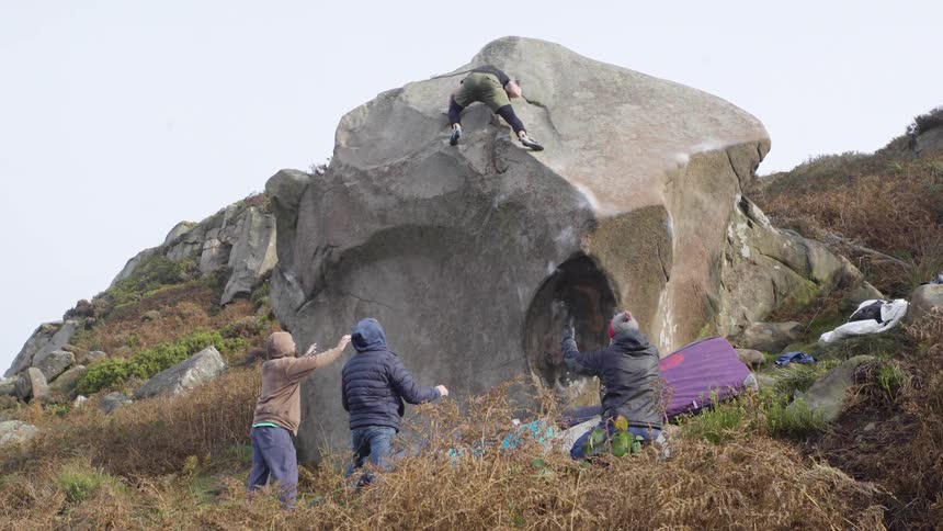 Climber ascending a large outdoor boulder while three people watch and provide support