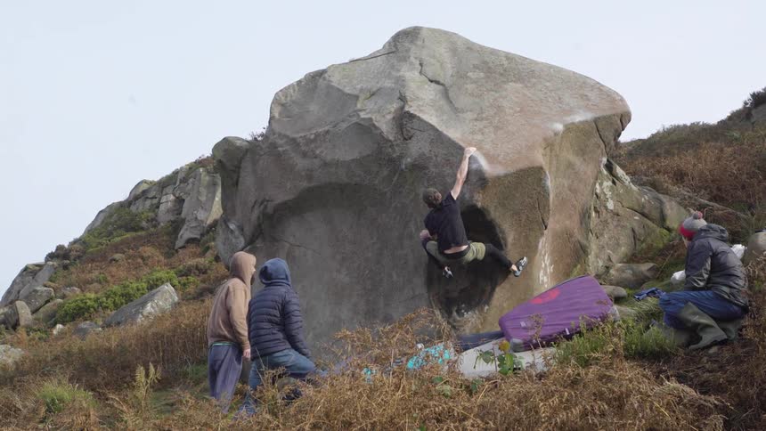A group of three young adult male friends all wearing winter jackets watch as one of them climbs a mountain at midday
