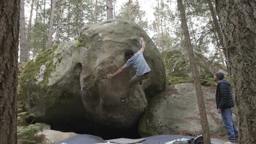 A man climbs a mountain in the woods at midday while another man stands nearby watching him
