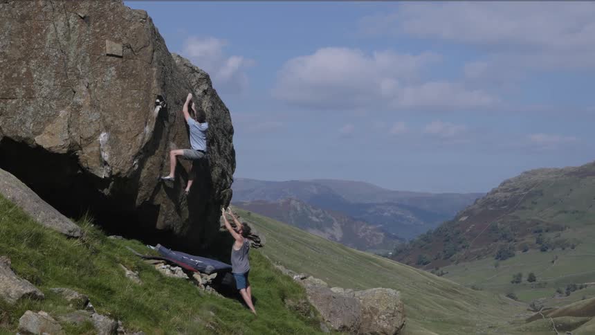 Man climbs boulder on grassy mountainside with a spotter nearby